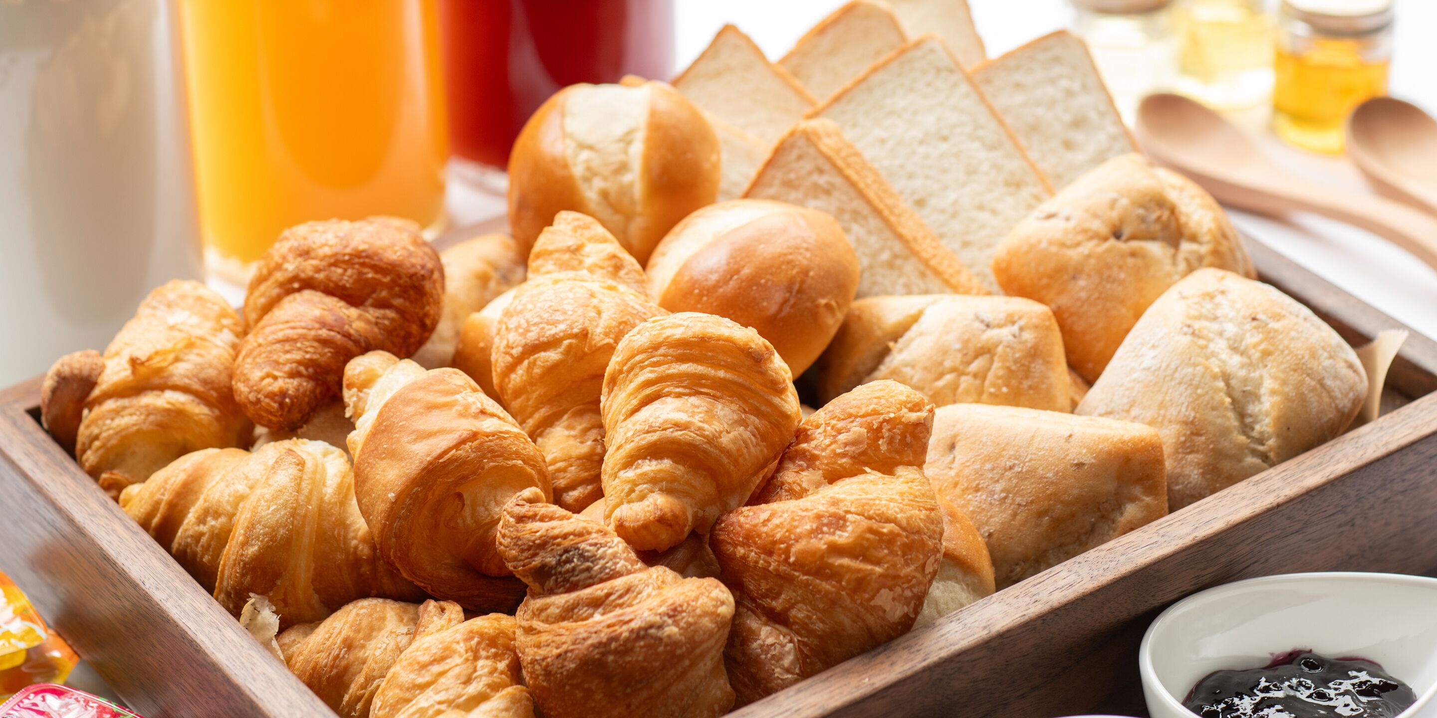 Een houten dienblad met verschillende soorten brood en gebak, waaronder croissants en stokbroden, met een klein schaaltje jam.