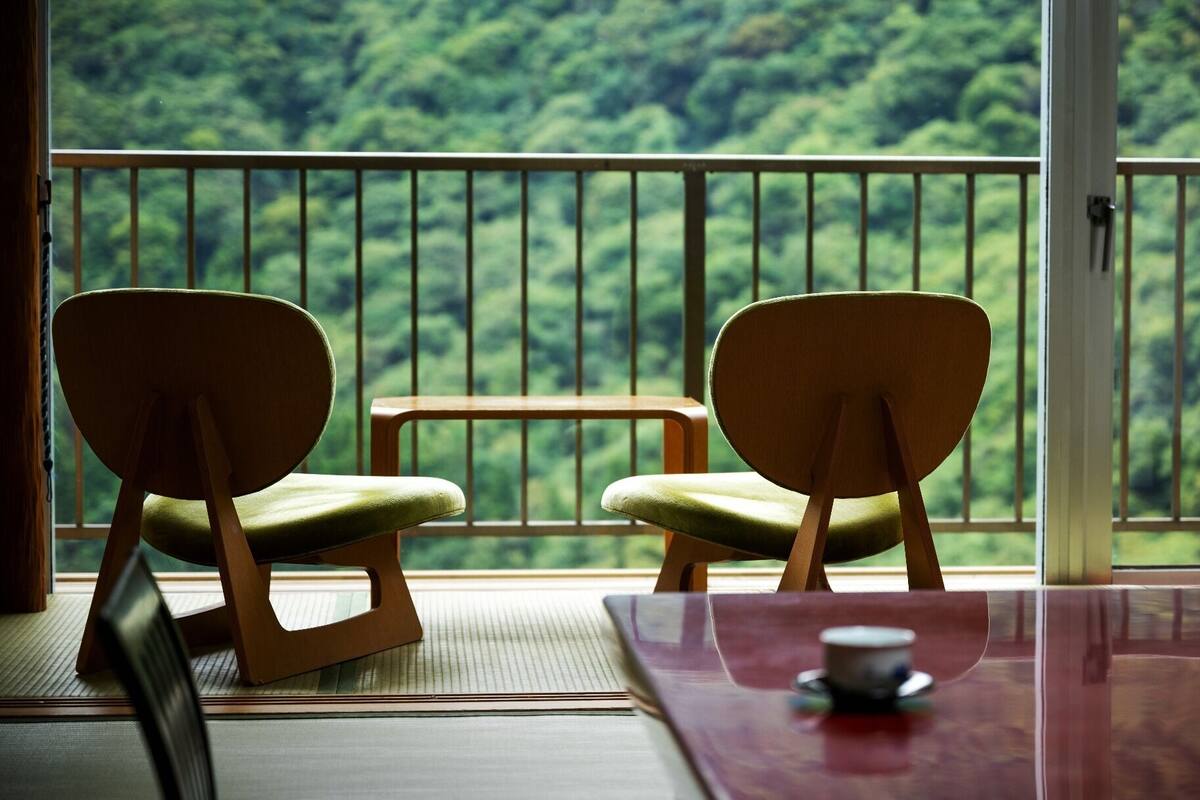 A room with two wooden chairs and a small table, a view of a forest through a balcony railing.
