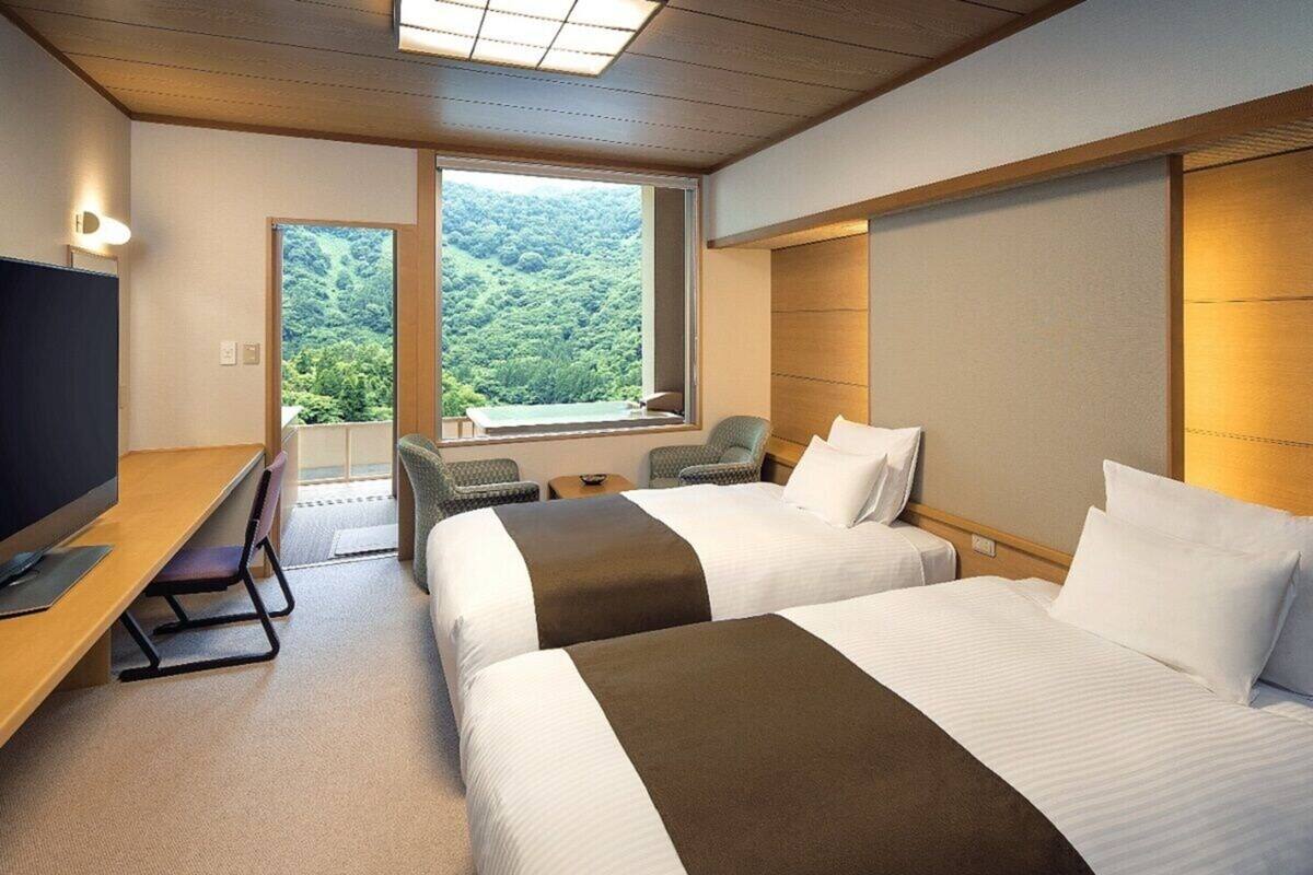 A hotel room with two beds, a desk, a TV, and a view of a forested area.