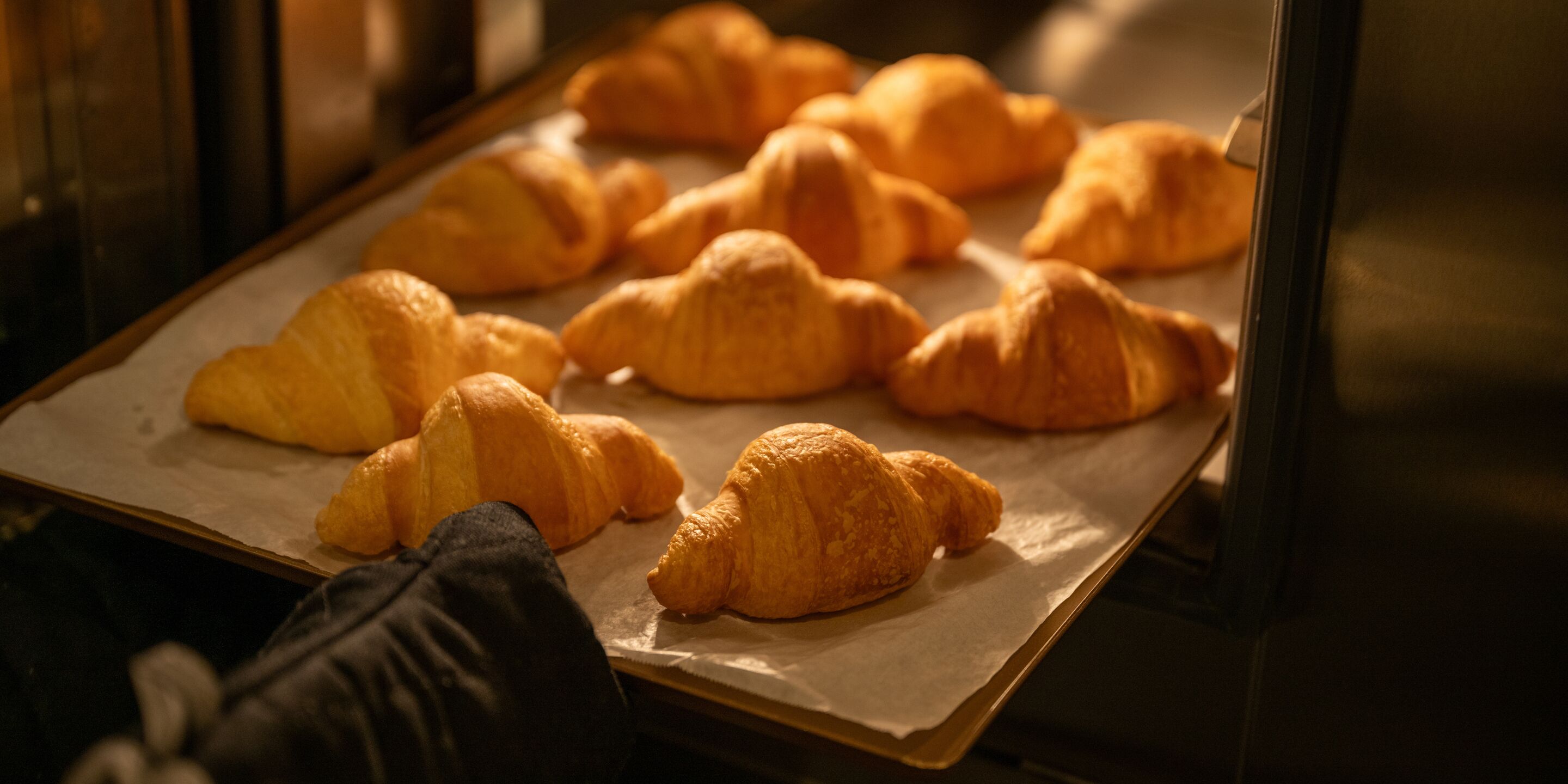 A tray of freshly baked croissants on parchment paper, likely in a bakery or kitchen setting.
