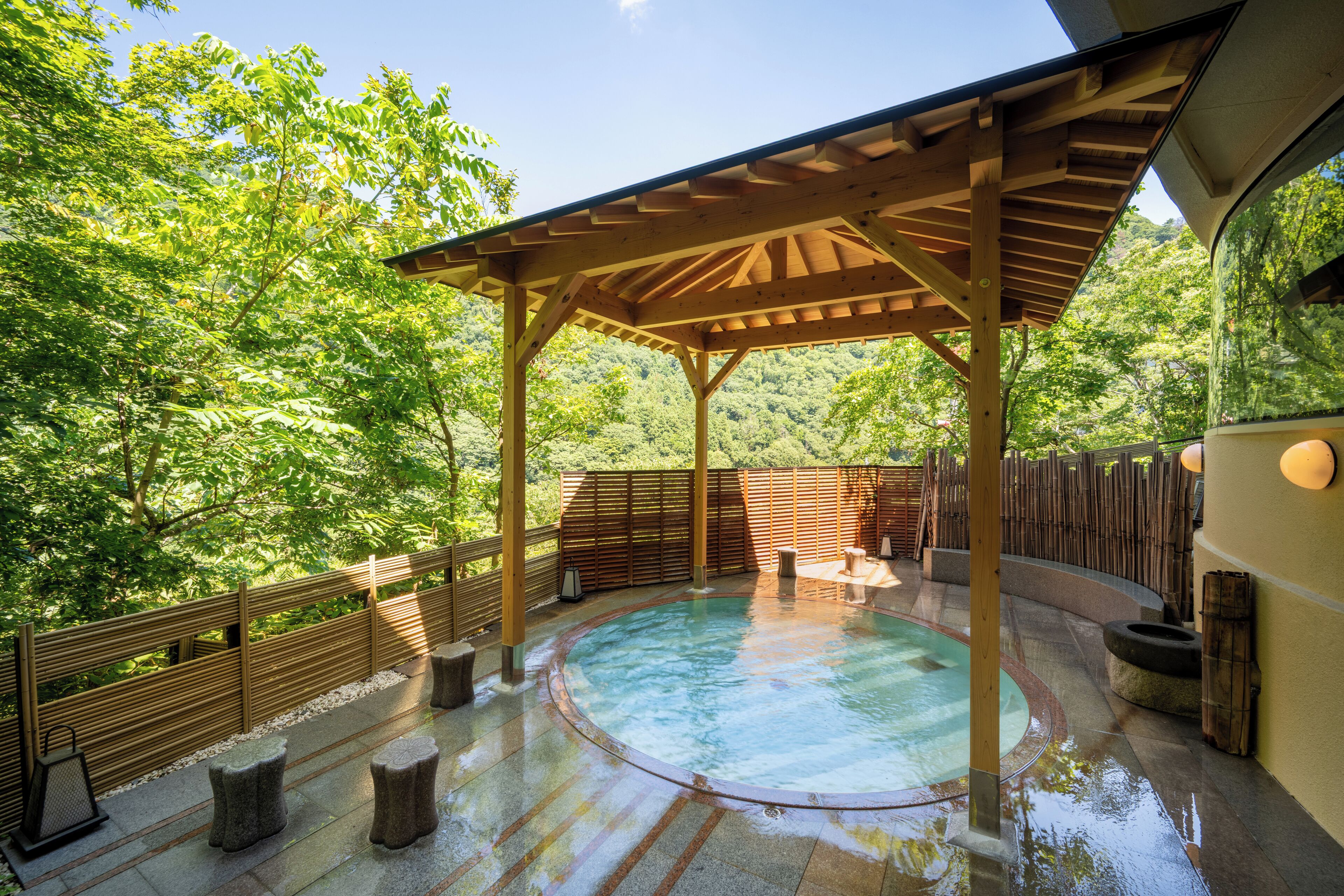A hot spring area with a wooden pergola, stone seating, and a pool surrounded by a wooden fence and lush greenery.