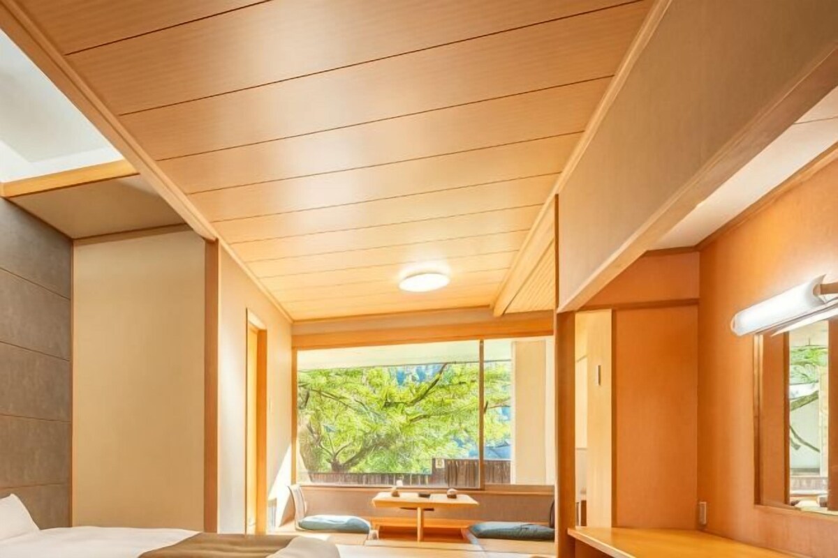 A hotel room with a wooden ceiling, a bed, a small table, and a window with a view of trees.