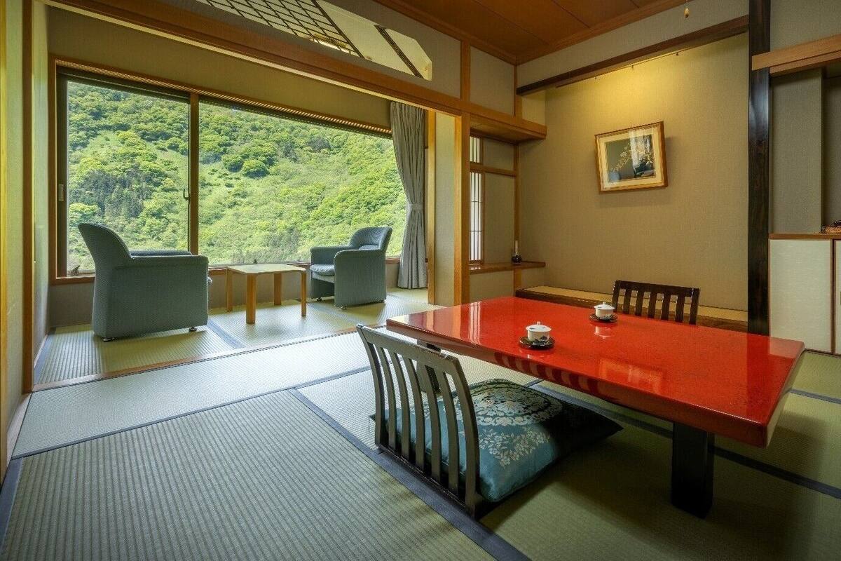 A traditional Japanese room with tatami flooring, a low table with cushions, and a view of a forested hillside.