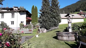 A white building with balconies, a tall coniferous tree, a wooden fence, and a fountain. - Apartment with garden in prime location (Tobadill)