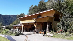 A wooden building with a covered porch, a driveway, and a mountainous backdrop. - Apartment with garden in prime location (Tobadill)