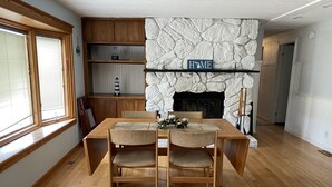 Uma sala de jantar com uma mesa e cadeiras de madeira, lareira de pedra e prateleiras de madeira.