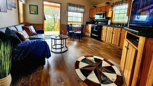 A cozy living room with a sofa, a coffee table, and a dining area with chairs. There's a kitchen with wooden cabinets and a TV mounted on the wall. - Cozy Canyon Lake Tiny House – Stunning Views & Great Location (Canyon Lake)