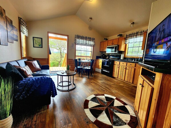 A cozy living room with a sofa, a coffee table, and a dining area with chairs. There's a kitchen with wooden cabinets and a TV mounted on the wall. - Cozy Canyon Lake Tiny House – Stunning Views & Great Location (Canyon Lake)
