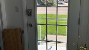 A white door with a grid pattern, a wooden door closer, and a small window with a view of a parking lot and greenery.