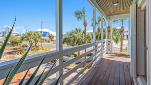 A wooden deck with a white railing, overlooking a beach with palm trees and a clear blue sky.