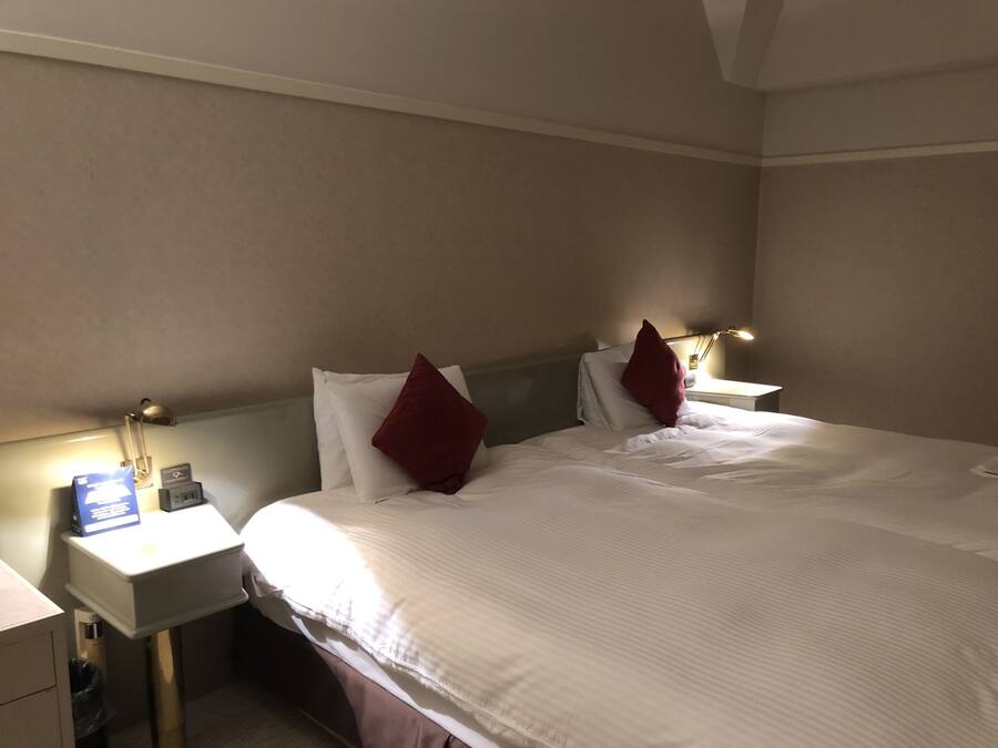 A hotel room with a bed, two bedside tables, a nightstand, a lamp, and a wall-mounted light.