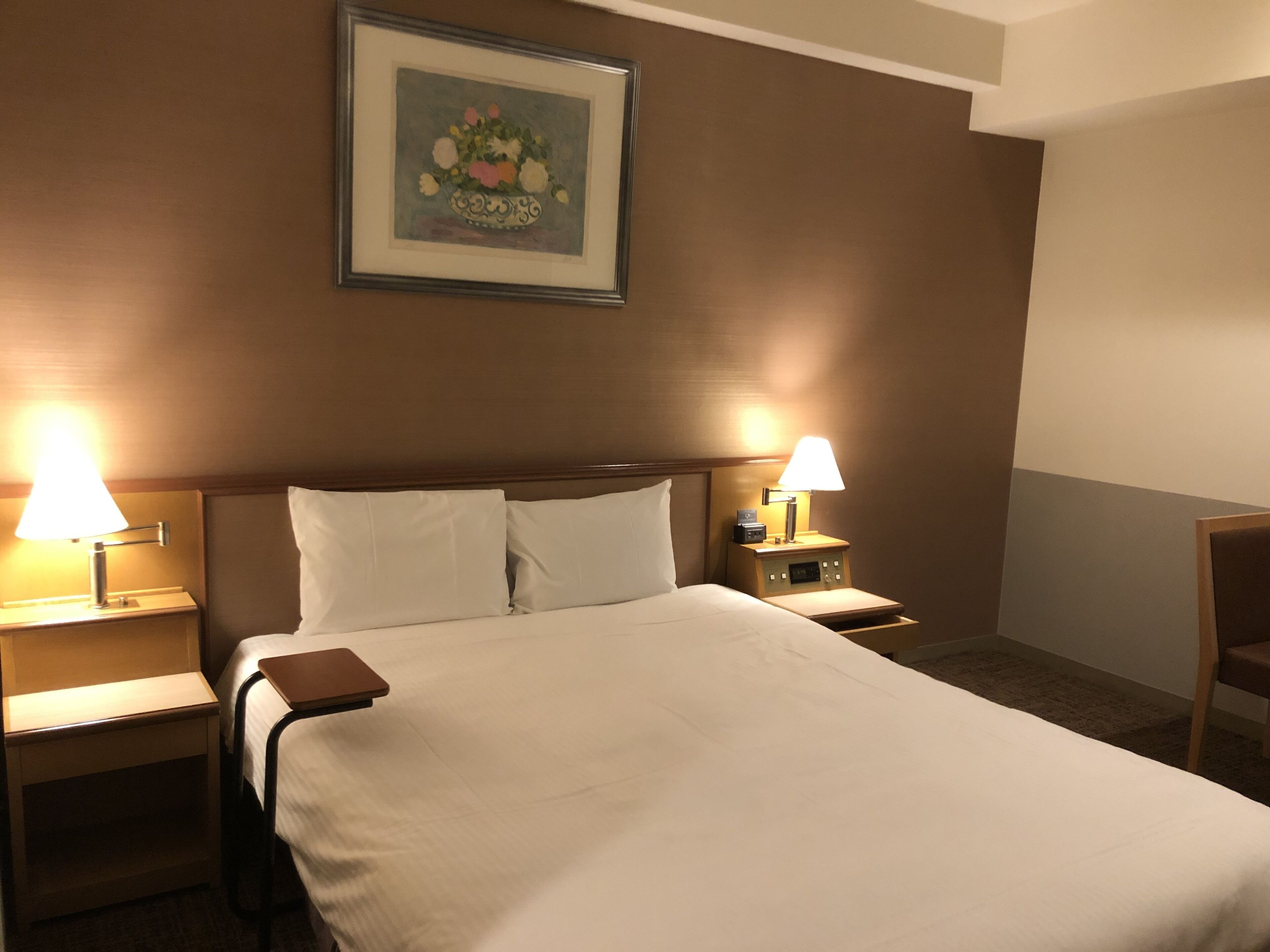 A hotel room with a large bed, two bedside tables with lamps, a framed floral painting on the wall, and a chair in the corner.