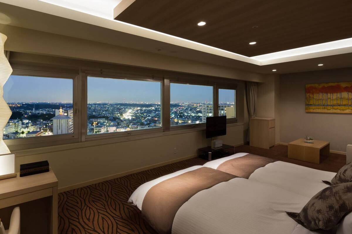 A hotel room with a large bed, a desk, a TV, and a view of the city at night.