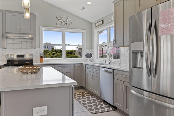 A modern kitchen with grey cabinetry, a stainless steel refrigerator, and a large window offering a view of the outside. - OCEAN VIEW, across from beach access, BEAUTIFULLY updated 4 bed,4 bath,sleeps 14 (Emerald Isle)