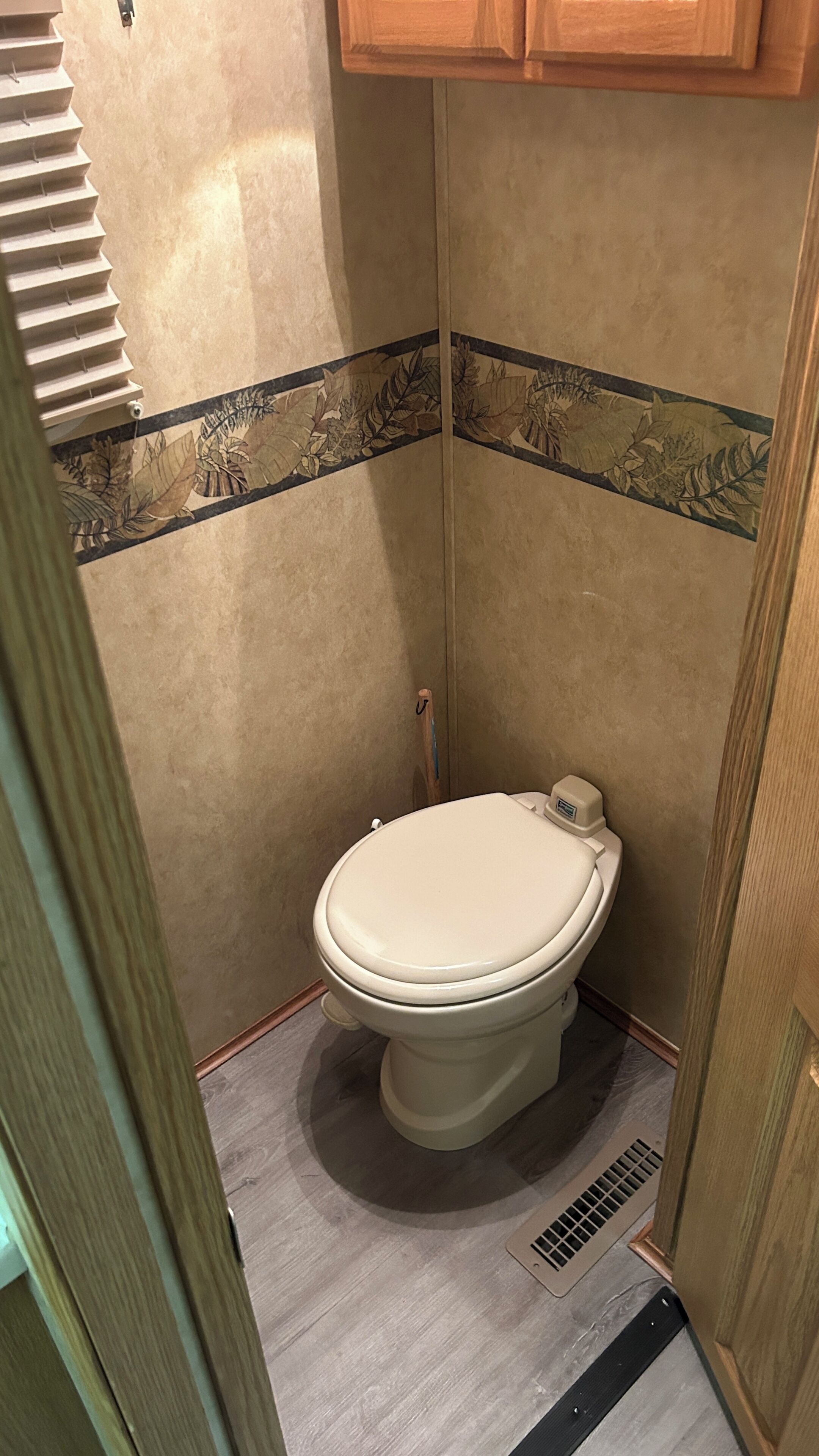 A small, compact bathroom with a toilet, a wooden cabinet above, and a vent on the floor.