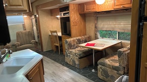 En kompakt campingvogns indretning med et tekøkken, et spiseområde med bord og stole, og et vindue med udsigt til grøn natur.