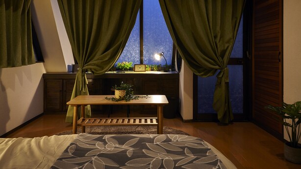 Une chambre avec une grande fenêtre, une table de chevet avec une lampe et un lit recouvert d’une couvre-lit à motifs floraux.