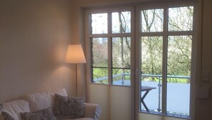 A room with a white sofa, a small white table, and a large window with a view of greenery outside. - Huus Windroos (Jemgum)