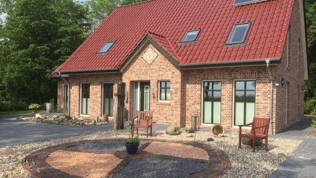 A brick house with a red roof, two skylights, and a circular gravel driveway with a fire pit. - Huus Windroos (Jemgum)