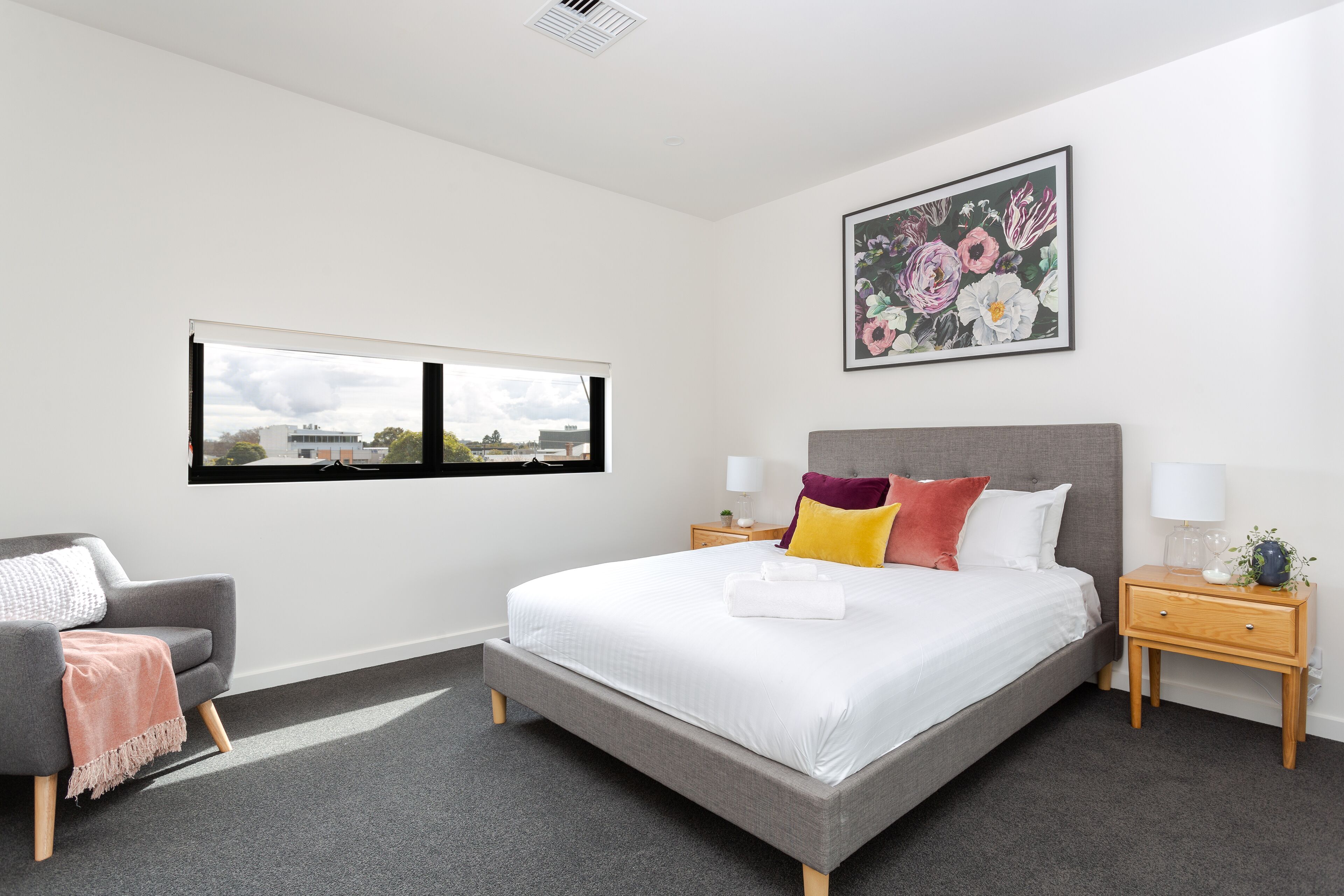 A modern bedroom with a large bed, a grey armchair, a wooden nightstand, a window with a view, and a floral artwork on the wall.