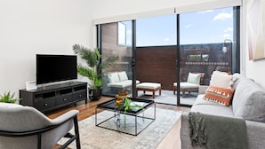 A modern living room with a flat-screen TV, a glass-top coffee table, a grey sofa, and a sliding glass door leading to an outdoor patio. - Little Grenfell Apartments by Urban Rest (Kent Town)