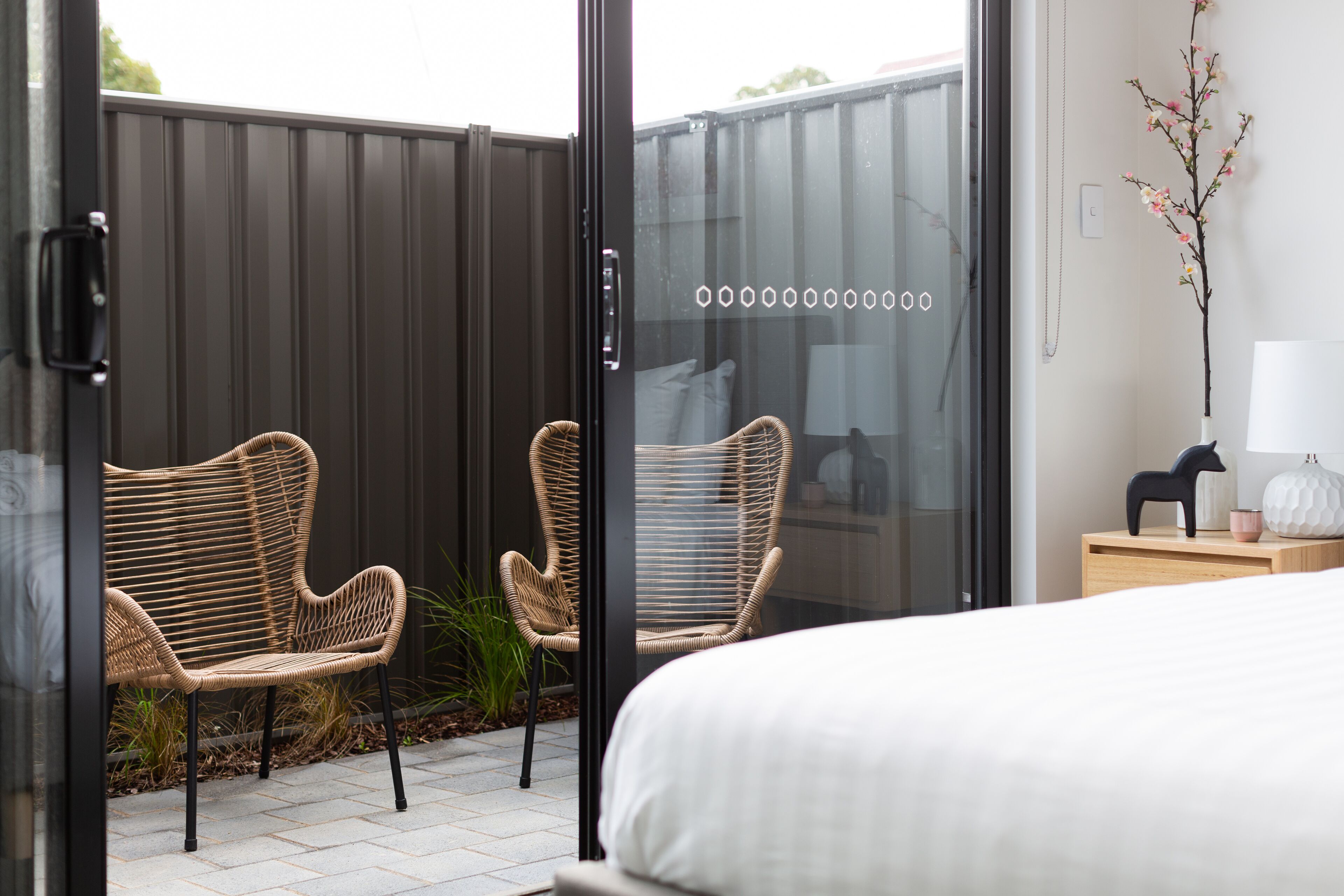 A modern bedroom with a large bed, two wicker chairs, a small wooden nightstand, and a vase with flowers.