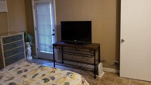 Un dormitorio con una cama, un televisor sobre un mueble bajo, un aparador, una puerta y un ventilador.