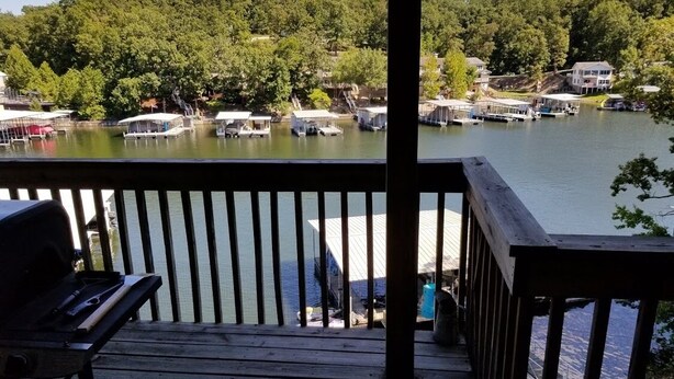 Una terraza de madera con una parrilla y vistas a un lago con barcos y casas al fondo.