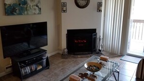 Una sala de estar con televisor de pantalla plana, chimenea, una mesa de centro de vidrio y un cuadro en la pared.