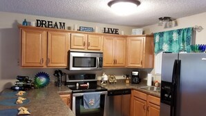Una cocina con armarios de madera, microondas, horno, lavavajillas y refrigerador de acero inoxidable. Hay objetos decorativos en la encimera y una toalla colgada en la puerta del horno.