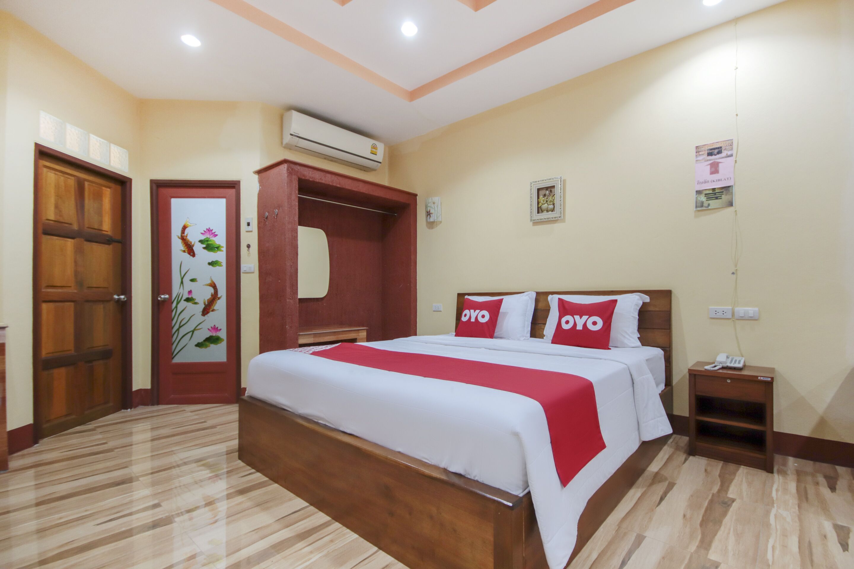 A hotel room with a large bed, wooden headboard, and a red and white bedspread. There is a wooden wardrobe, a small bedside table with a phone, and a wall-mounted air conditioning unit.