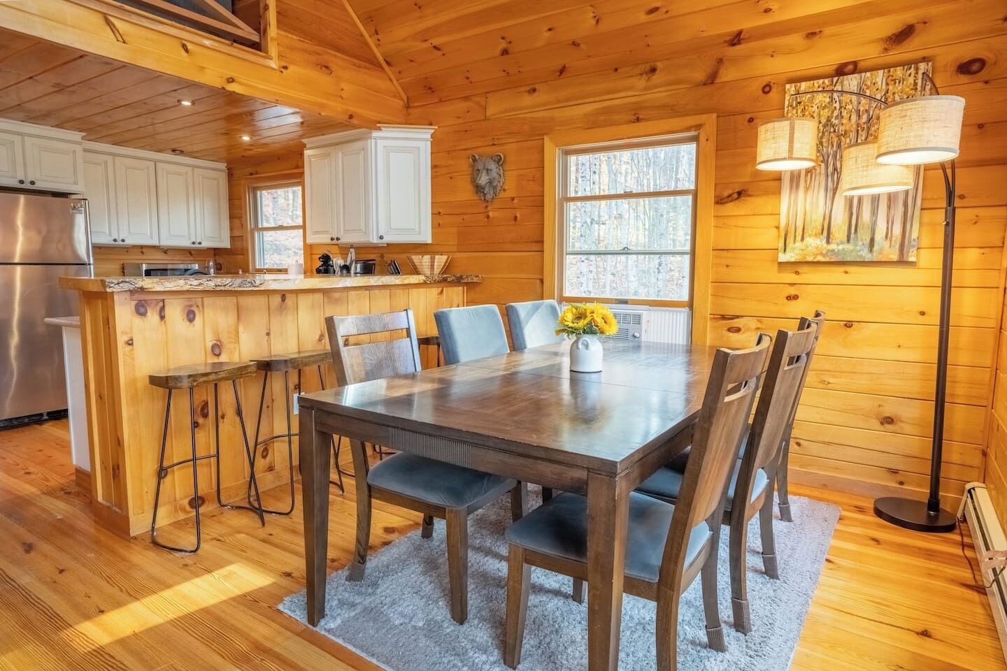 Interior de una cabaña de madera con una mesa de comedor, sillas, una zona de cocina y un ventanal grande.