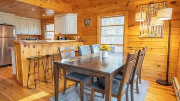 Interior de una cabaña de madera con una mesa de comedor, sillas, una zona de cocina y un ventanal grande.
