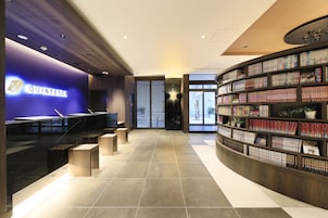 Eine moderne Hotellobby mit einem 'QUINTESSA'-Schild, einer Rezeption, einem großen Bücherregal und einem Fliesenboden.
