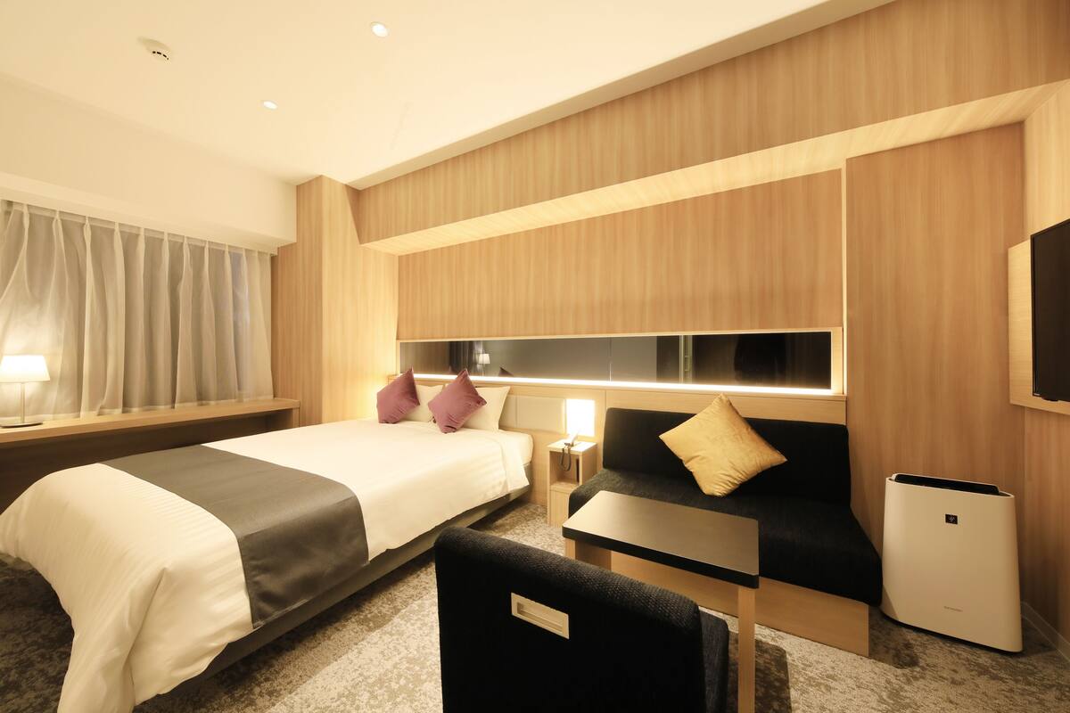 A hotel room with a bed, a sofa, a desk, and a trash bin.