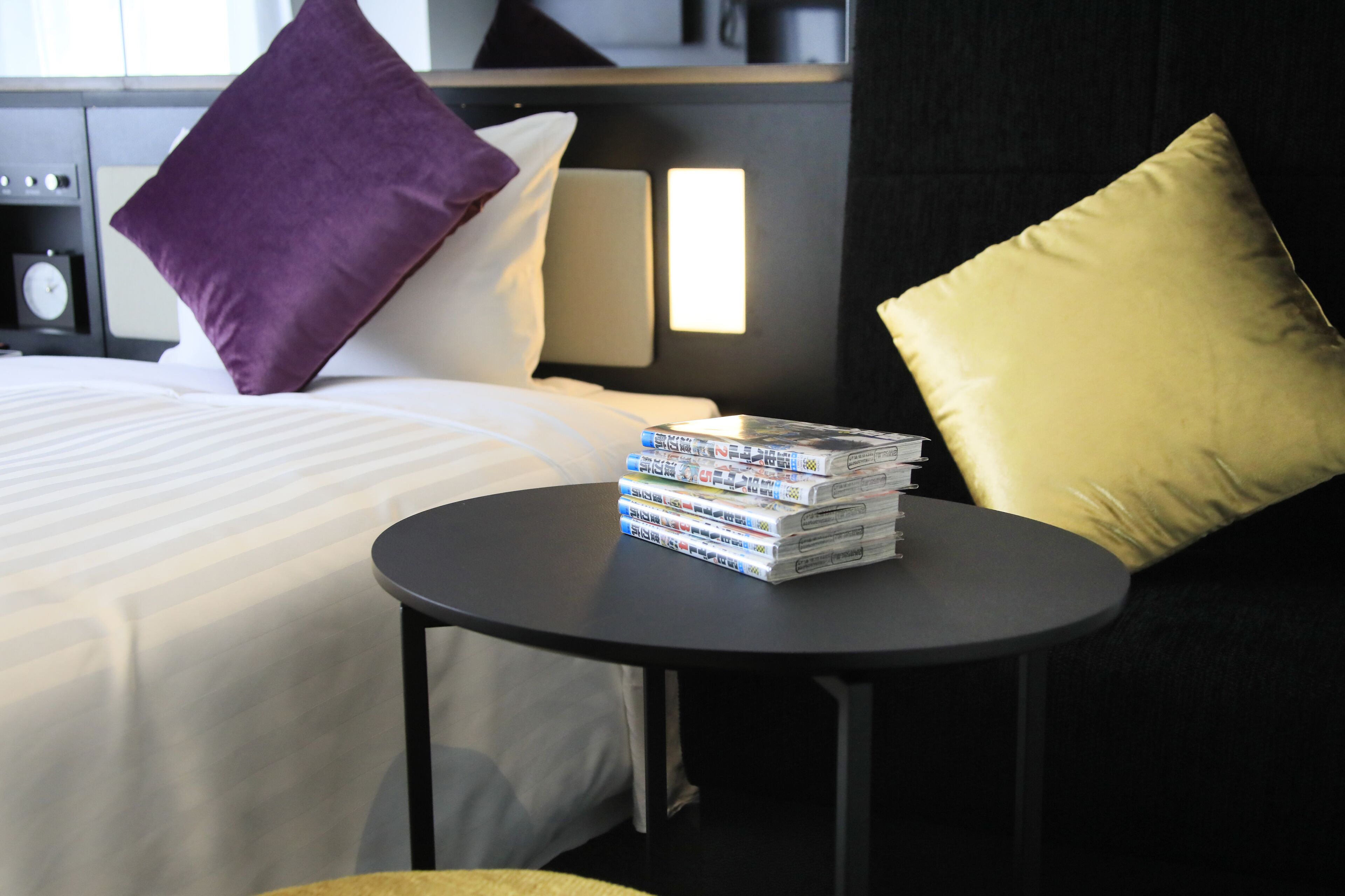 A neatly made bed with white linens, a stack of magazines on a small round table, and two pillows, one purple and one yellow, on the bed.
