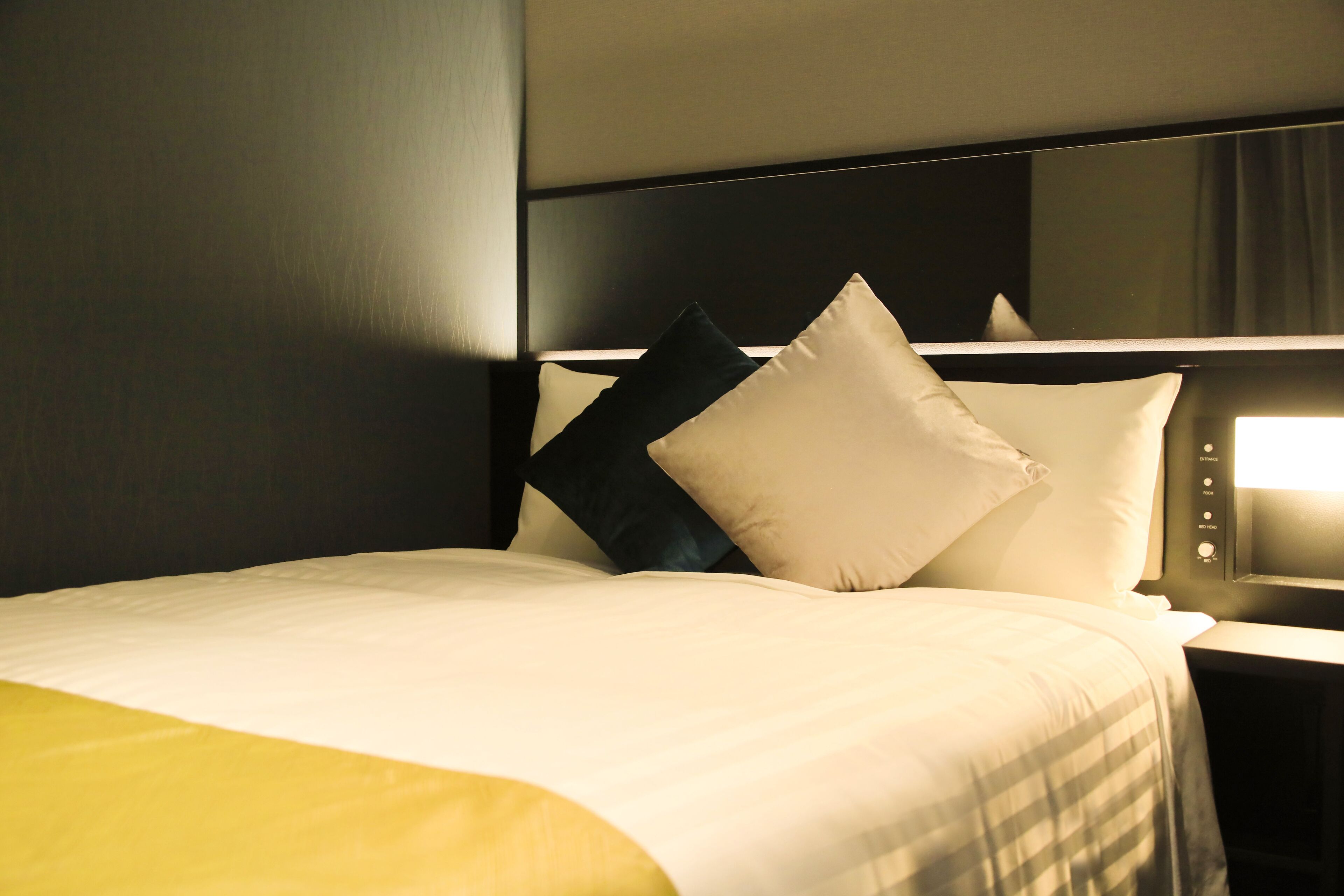 Un lit bien fait, recouvert d’une couvre-lit blanche et jaune, avec une tête de lit et une table de chevet surmontée d’une lampe.
