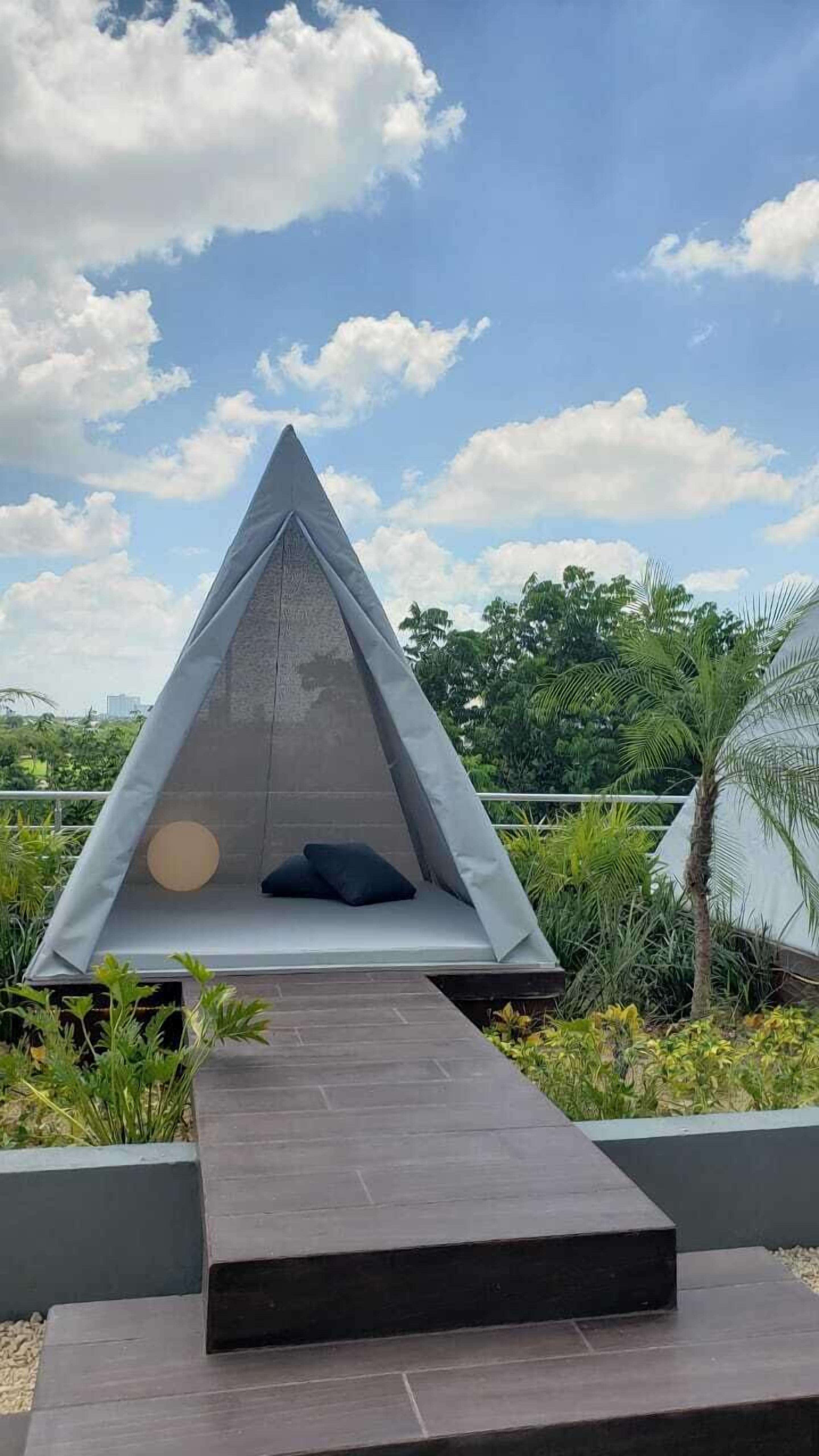 A modern, triangular outdoor seating area with a bench and cushions, surrounded by lush greenery and a clear sky.