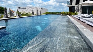Une piscine avec des chaises longues et des parasols, un bâtiment moderne orné d’une enseigne « Liverpool », et un ciel dégagé.