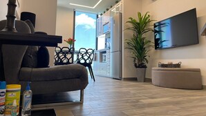 Un salon moderne avec un canapé, une table basse et un téléviseur fixé au mur. On aperçoit, à l’arrière-plan, une plante en pot et un coin repas.