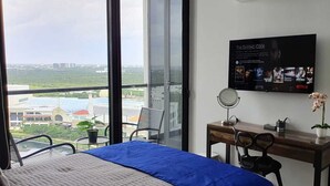 Une chambre d’hôtel dotée d’une grande fenêtre donnant sur la ville, d’un lit recouvert d’une courtepointe bleue, d’un bureau avec un miroir et d’une orchidée en pot.