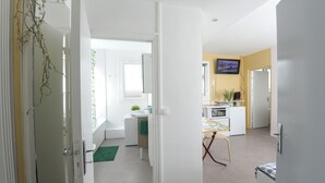 A modern kitchen with white appliances, a dining area with a table and chairs, and a view of the living room through an open door. - Studio Cosy et Fonctionnel, Proche de Paris (Villeneuve-la-Garenne)
