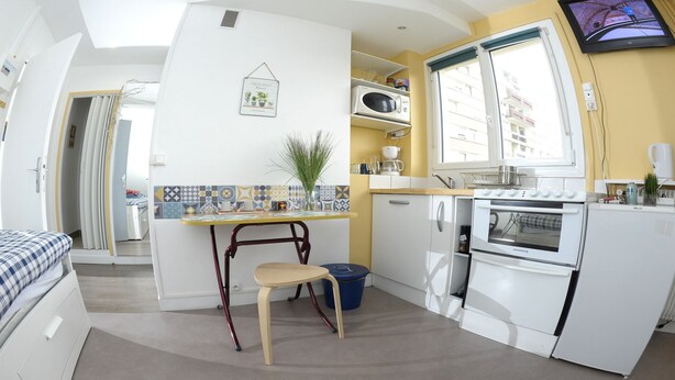 A compact kitchen with a microwave, oven, and sink. There's a small dining table with a chair, a blue trash bin, and a potted plant. - Studio Cosy et Fonctionnel, Proche de Paris (Villeneuve-la-Garenne)