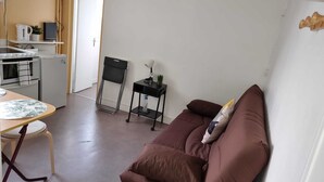 A compact living space with a kitchenette, a dining area with a table and chairs, a sofa with a cushion, and a small side table with a lamp and a plant. - Studio Cosy et Fonctionnel, Proche de Paris (Villeneuve-la-Garenne)