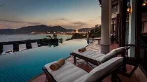 Uma área à beira da piscina com espreguiçadeiras e vista para uma cidade costeira ao entardecer.