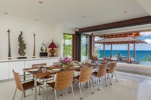 Un comedor moderno con vistas al océano, que incluye una mesa y sillas de madera, adornos en la pared y un ventanal que da a una zona de estar al aire libre.