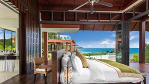 A bedroom with a large bed, a chair, a bedside table, and a view of the ocean. - Baan Santisuk 5 Bedroom (Kamala)