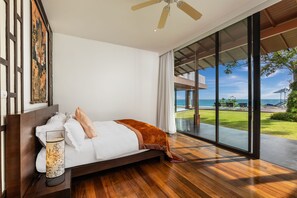 Un dormitorio moderno con una cama grande, suelo de madera, ventilador de techo y vistas al océano a través de ventanales.