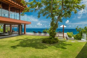 Un espacio exterior moderno con un salón envuelto en vidrio, mobiliario para exteriores y vista al océano.