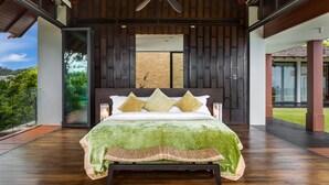 A bedroom with a large bed, wooden flooring, a ceiling fan, and a view of greenery outside. - Baan Santisuk 5 Bedroom (Kamala)